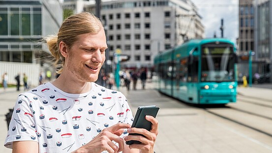 Eine Person tippt auf ihrem Handy, im Hintergrund fährt eine Straßenbahn in die Haltestelle Willy-Brandt-Platz ein.