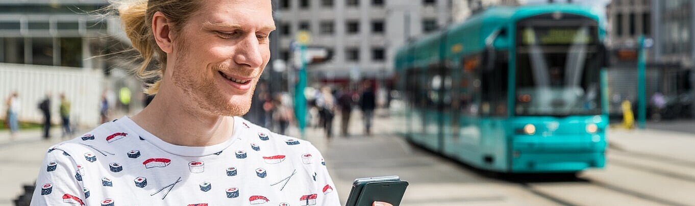 Eine Person tippt auf ihrem Handy, im Hintergrund fährt eine Straßenbahn in die Haltestelle Willy-Brandt-Platz ein.