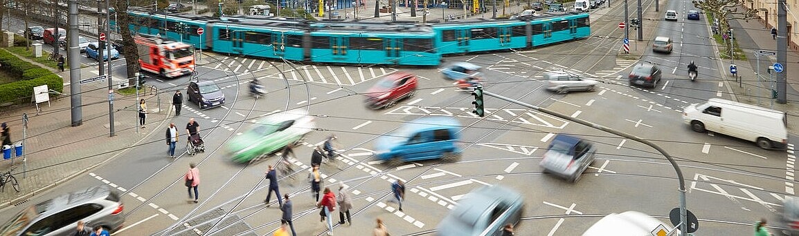 Eine große Straßenkreuzung mit Ampeln auf der Fußgänger, Autos und eine Straßenbahn gleichzeitig unterwegs sind.