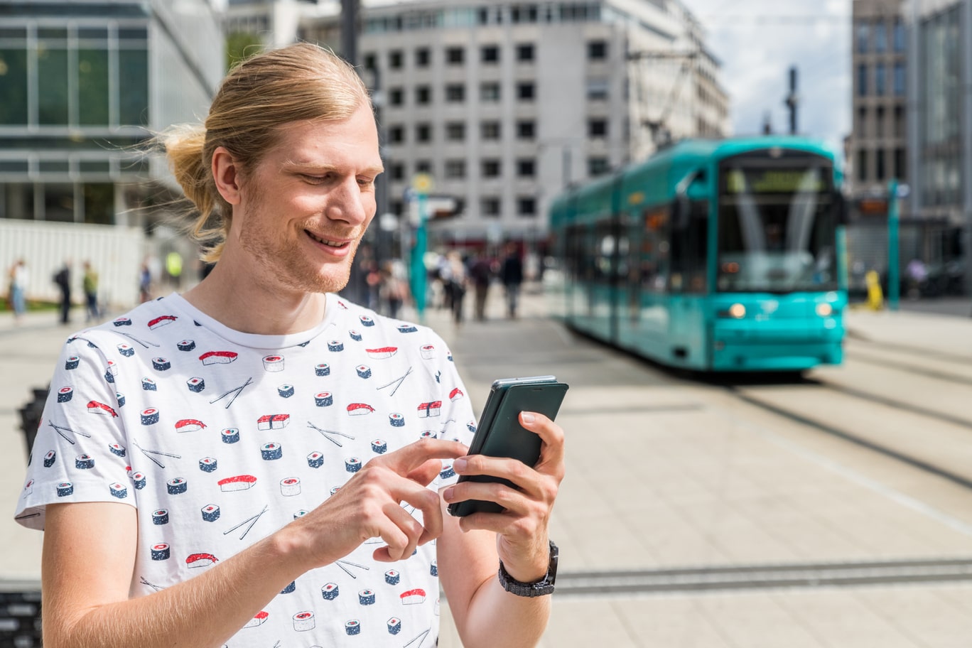 Eine Person tippt auf ihrem Handy, im Hintergrund fährt eine Straßenbahn in die Haltestelle Willy-Brandt-Platz ein.